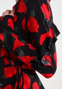 Sort blomsterkjole med røde roser, der har flæseærmer og en bindebånd i taljen. Stoffet er let og struktureret. Grønne blade fremhæver blomsterne.