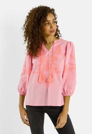 Mujer con blusa rosa con bordado floral y lazos con flecos, combinada con vaqueros oscuros, de pie frente a un fondo claro y liso.