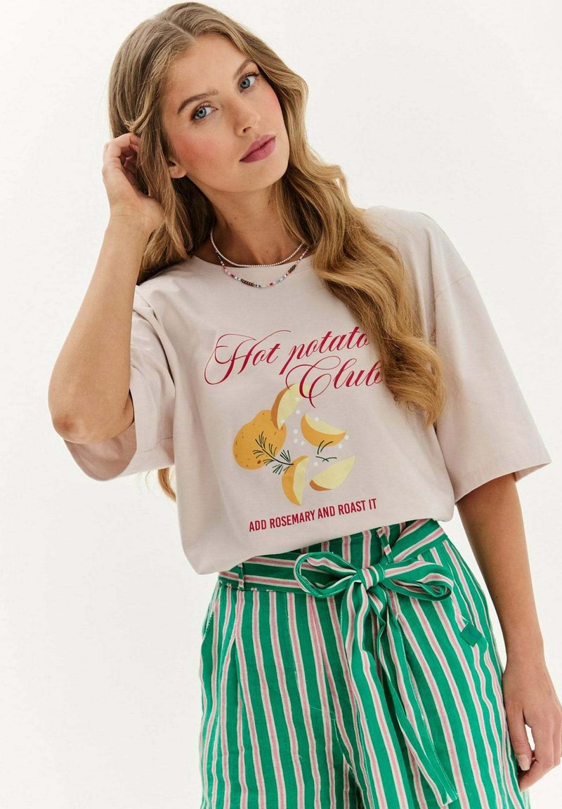 Mladá žena s dlouhými blond vlasy, oblečená v lehké béžové košili s nápisem "Hot potato Club" a zelenými pruhovanými kalhotami s uvázanou mašlí v pase.