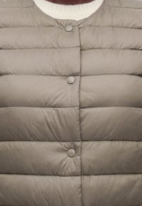 Veste matelassée marron clair avec coutures horizontales et boutons-pression sur un pull blanc à col rond côtelé.