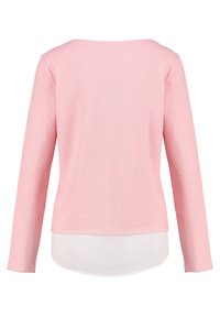 Vaaleanpunainen pitkähihainen paita, jossa on tekstuurikuvio, pyöreä pääntie ja hieman pidempi takasauma, kerroksittain valkoisen aluspaidan päällä.