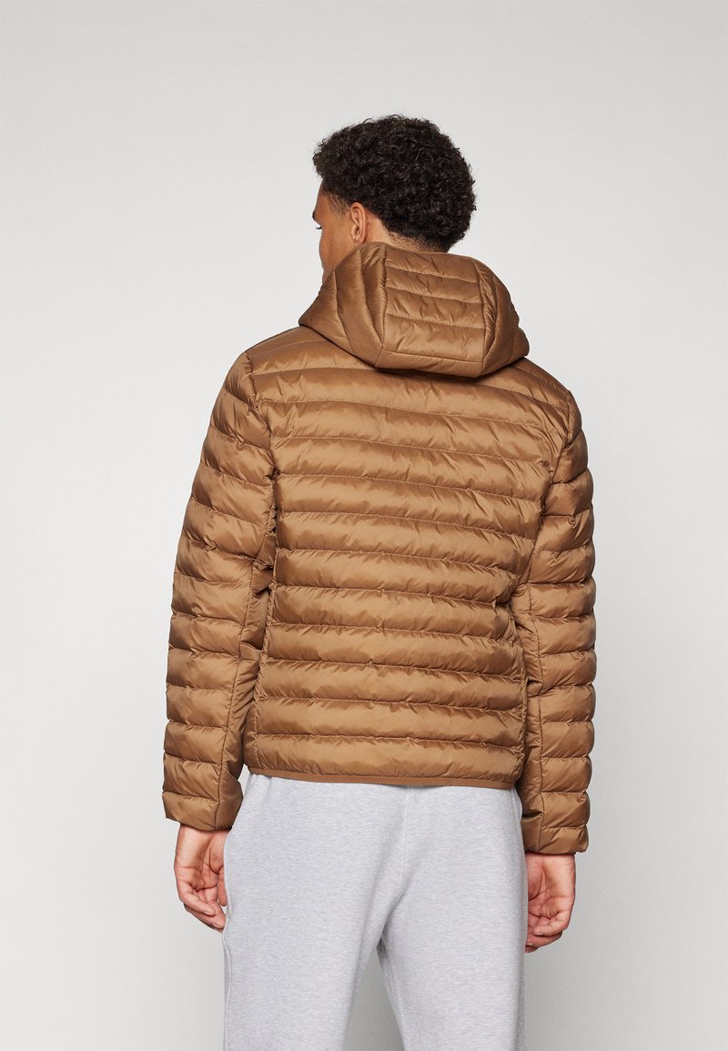 Giacca a vento marrone con motivo a righe orizzontali, cappuccio integrato e texture liscia. Indossata sopra dei pantaloni della tuta grigi, che mostrano un fit rilassato.