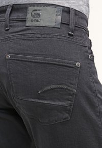 Jeans de mezclilla negros con tejido texturizado, diseño de cinco bolsillos y una pieza rectangular de cuero con logo en relieve en la parte trasera.