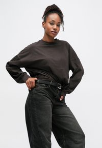 Σκούρο καφέ φαρδύ φούτερ με χαμηλούς ώμους, συνδυασμένο με άνετα μαύρα τζιν και μαύρη ζώνη. Ομαλό ύφασμα με ελάχιστη υφή.