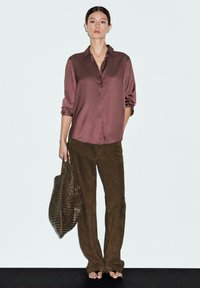 Camicia di raso marrone con bottoni frontali, maniche arrotolate; abbinata a pantaloni scamosciati marroni; tiene una borsa di paglia marrone.