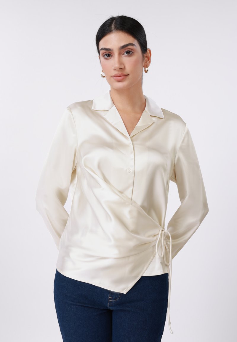 Μπλούζα από σατέν κρέπα με γιακά, σχεδιασμό τύπου wrap, κουμπιά στο μπρο στινό μέρος και διακοσμητικό κορδόνι στη μέση. Συνδυάζεται με σκούρο μπλε τζιν παντελόνι.