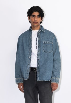 Ung mand med mørkt krøllet hår iført en åben blå denimjakke over en hvid skjorte og sorte bukser mod en enkel lys baggrund.