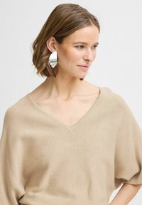 Top en tricot beige avec un décolleté en V et des manches larges. Le mannequin porte de grandes boucles d'oreilles géométriques en argent. Texture lisse, design décontracté.