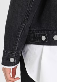 Rękaw czarnej kurtki dżinsowej z srebrnymi guzikami; widoczne przeszycia i kontrastowy biały brzeg koszuli pod spodem. Gładka faktura.