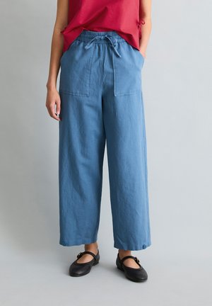 Frau trägt lockere blaue Leinenhose mit Taschen vorne und Knopfdetails an den Säumen, kombiniert mit schwarzen Mary-Jane-Schuhen.