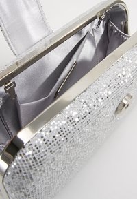 Pochette scintillante argentée avec cadre en métal. L'intérieur présente une doublure grise lisse et des compartiments pour l'organisation. Détail de fermeture sécurisé.