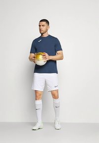 Vīrietis tumši zilā kreklā, baltās šortās un zeķēs tur dzeltenu un baltu futbola bumbu, stāvot pret vienkrāsainu gaišu fonu.