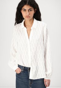 Camisa blanca de botones con un patrón de diamantes texturizado, mangas largas y cuello con muesca, combinada con una falda de mezclilla.