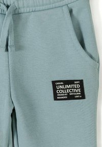 Pantalon de survêtement bleu clair avec taille à cordon et poche avant, avec un écusson noir marqué "Unlimited Collective" en texte blanc.