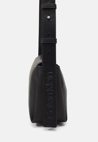 Schwarze Leder-Schultertasche mit glatter Textur, geprägtem Logo am Träger und verstellbarem Schnallenverschluss für die Länge. Kompakte, rechteckige Form.