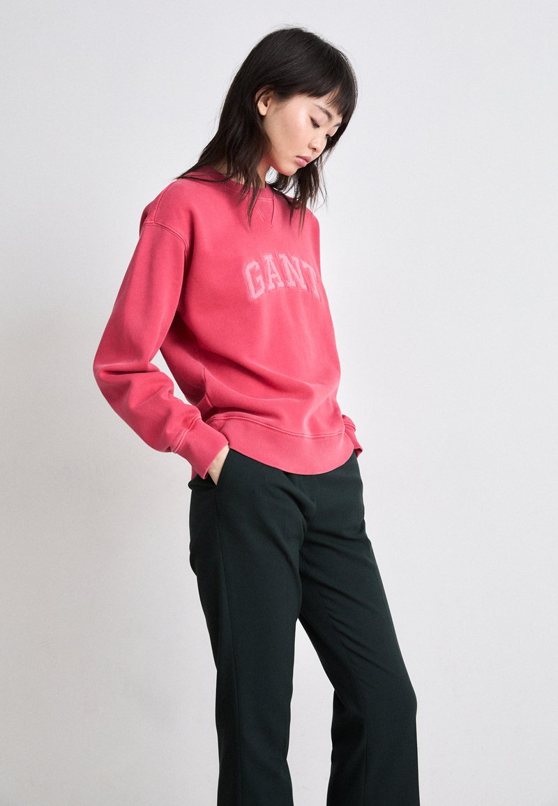 Różowa bluza z kapturem z logo "GANT", ściągacze na rękawach i u dołu, zestawiona z czarnymi spodniami. Miękki materiał i luźny krój, z okrągłym dekoltem.