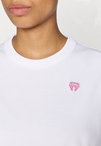 Bijela pamučna majica kratkih rukava s okruglim rubom vrata i malim ružičastim izvezenim logotipom na prsima. Glatka tekstura s udobnim krojem za ležeran izgled.