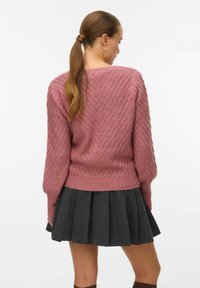 Roosa kootud sviiter, millel on tekstuuriline ševroni muster, koos tumeda halli volditud seelikuga. Riietusel on pikad varrukad ja kinnitatud talje.