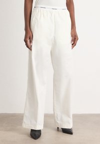 Calvin Klein white cotton blend trousers with an elastic waistband and wide-leg fit, worn with black pointed heels. Smooth texture.