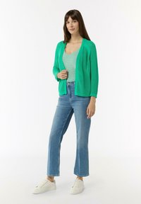 Grüne, offen gestrickte Strickjacke über einem gestreiften Oberteil, kombiniert mit hellblauer Jeans und weißen Sneakers. Mit langen Ärmeln und einer lockeren Passform.