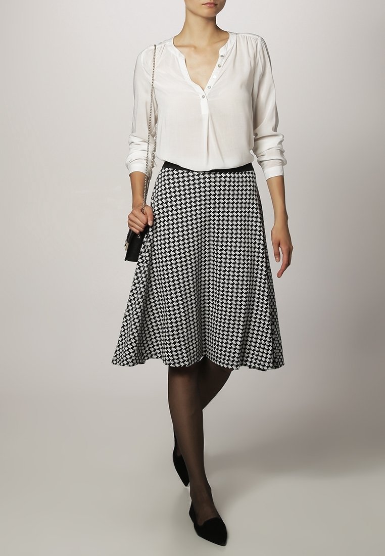 Femme portant un chemisier blanc à manches longues, une jupe pied-de-poule noire et blanche, des collants noirs, des ballerines noires, et tenant un petit sac à main noir.