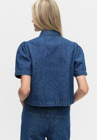 Een vrouw met blond haar draagt een blauwe denim overhemd met korte mouwen met een subtiel geborduurd patroon en bijpassende denim broek, met haar handen in de achterzakken.