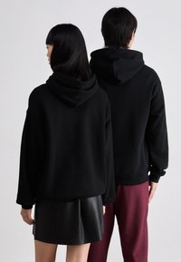 Sudaderas con capucha negras de corte relajado, con puños y cinturilla de canalé. La figura masculina lleva pantalones color burdeos, mientras que la figura femenina viste una falda de cuero negra.