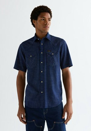 Młody mężczyzna w granatowej kratkowanej koszuli z krótkim rękawem zapinanej na guziki i ciemnych dżinsach, patrzący lekko z boku.