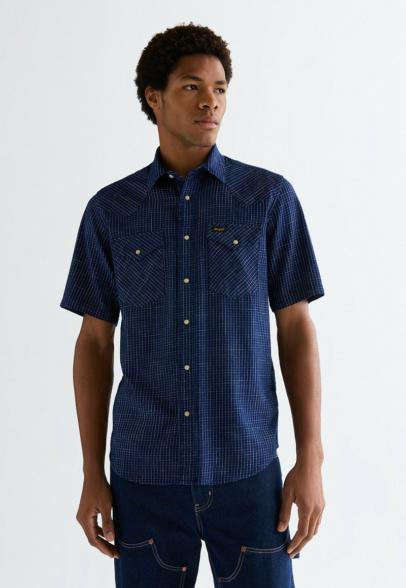 Giovane uomo che indossa una camicia a maniche corte a quadri blu navy e jeans scuri, che guarda leggermente di lato.