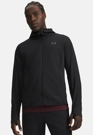 Man draagt een zwarte capuchonjas met rits, duimgaten en een subtiel logo op de borst, gecombineerd met een donkere broek en een bordeauxrood hemd eronder.