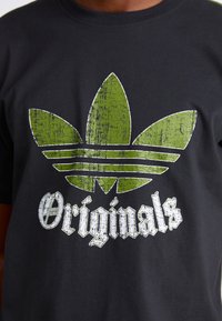 Fekete póló, nagy, zöld, repedezett textúrájú trefoil logóval, alatta fehér gotikus stílusú betűkkel az "Originals" felirattal.