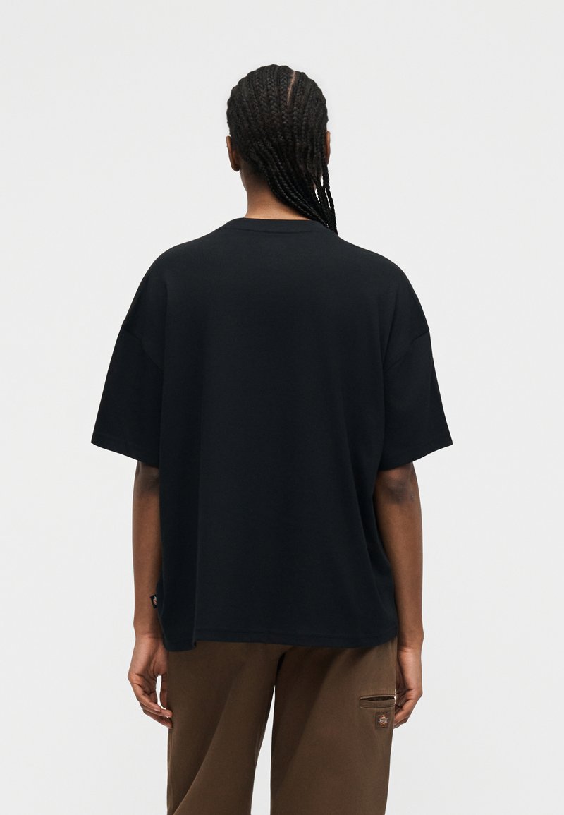 Persona con i capelli intrecciati vista di schiena, indossa una maglietta nera a maniche corte e larghe e pantaloni marroni con una tasca laterale.