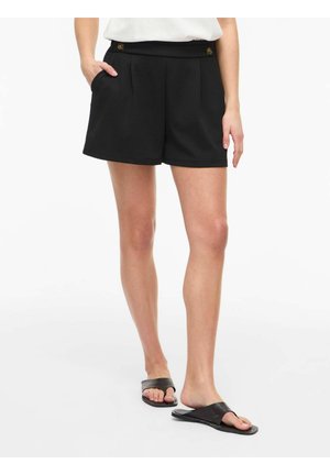 Nő fekete magasderekú rövidnadrágot visel elülső redőkkel és gombokkal, fekete papuccsal, fehér háttér előtt áll.