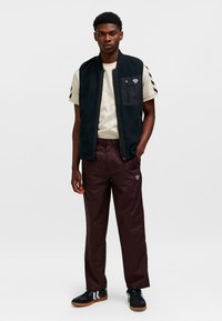 Zwarte fleecevest met een ritszak, beige T-shirt eronder, wijnrode broek en zwarte schoenen met witte strepen en een bruine zool.