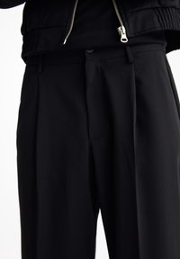 Gros plan sur une personne portant un pantalon noir sur mesure avec un bouton et des passants de ceinture, ainsi qu'une veste noire partiellement zippée.