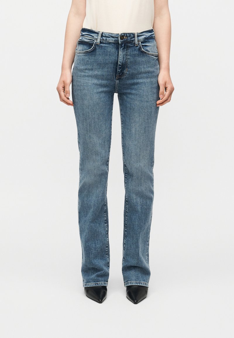 Μπροστινή όψη ατόμου που φορά μεσαίας μέσης ευθείς μπλε τζιν, συνδυασμένα με μαύρα παπούτσια μυτερά και λευκό αμάνικο τοπ.