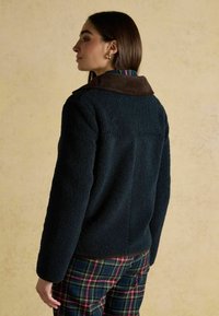 Giacca sherpa blu marina con colletto in corduroy marrone; presenta maniche dritti e una superficie testurizzata. Abbinata a pantaloni a quadri.