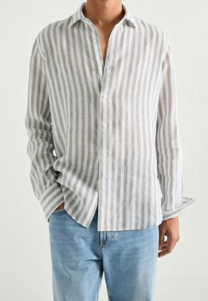 Homme portant une chemise à rayures verticales gris clair et blanc avec un jean bleu clair, une main dans la poche, sur un fond uni.