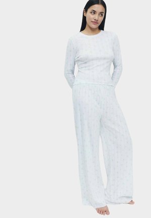 Donna che indossa una maglia bianca a maniche lunghe e pantaloni a gamba larga con sottili righe verticali e piccoli motivi verdi, in piedi scalza con le mani dietro la schiena.