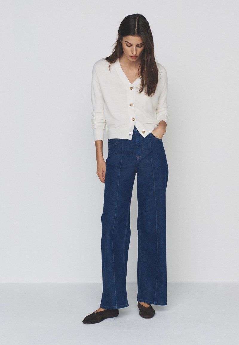 Woman wearing a white buttoned cardigan, high-waisted wide-leg blue jeans, and black flats, standing with one hand in pocket.