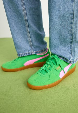 Personne portant un jean bleu et des baskets Puma vert vif et rose, debout sur un tapis vert clair.