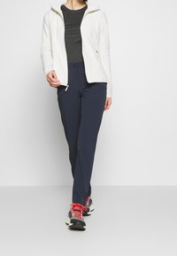 Frau, die auf einem weißen Hintergrund spaziert und ein schwarzes T-Shirt, eine weiße Zip-Up-Jacke, dunkelblaue Hosen und schwarze Laufschuhe mit roten Schnürsenkeln trägt.
