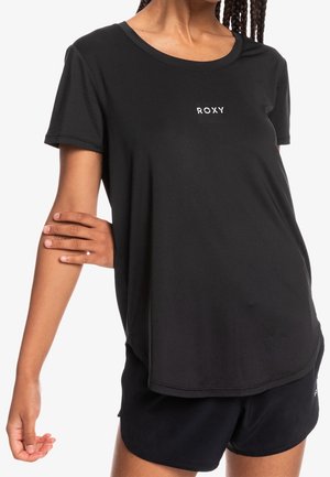 Kobieta ubrana w czarny, krótkim rękawem t-shirt Roxy i czarne spodenki, ręka zgięta z dłonią chwytającą przeciwną rękę.