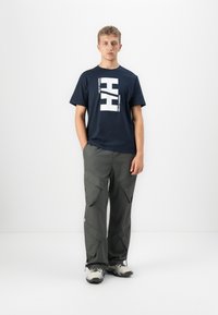 Junger Mann steht vor weißem Hintergrund, trägt ein dunkelblaues T-Shirt mit großem weißen "HH Helly Hansen"-Logo, graue Cargo-Hosen und Wanderschuhe.