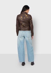 Veste en cuir marron ornée d'appliqués en forme d'étoile dorée, associée à un jean large bleu clair. La veste possède une coupe ajustée et des poignets zippés.