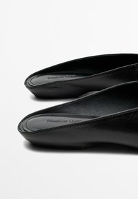 Zwarte leren instapplatte schoenen met een licht puntige neus en een zichtbaar merknaam die aan de binnenkant van de hiel is geperst.