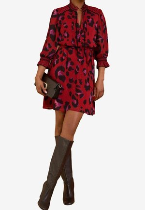 Kvinde iført en rød og lilla leopardmønstret kjole med lange ærmer, der holder en sort clutch og har knæhøje sorte støvler på.