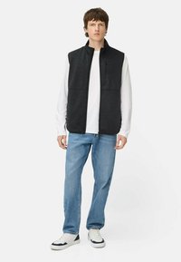 Graue Fleece-Weste mit hohem Kragen, Frontreißverschluss und zwei Seiten Taschen. Getragen über einem weißen Langarmshirt und kombiniert mit blauen Jeans.