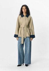 Beige jasje met riem en een grote marineblauwe kraag, brede mouwen en een knoopsluiting, gecombineerd met hoge blauwe denim en zwarte hakken.