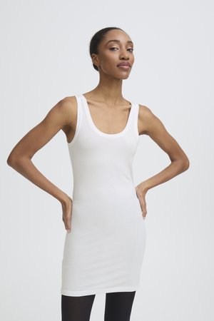 Femme en robe blanche ajustée sans manches, mains sur les hanches, debout contre un fond clair uni.